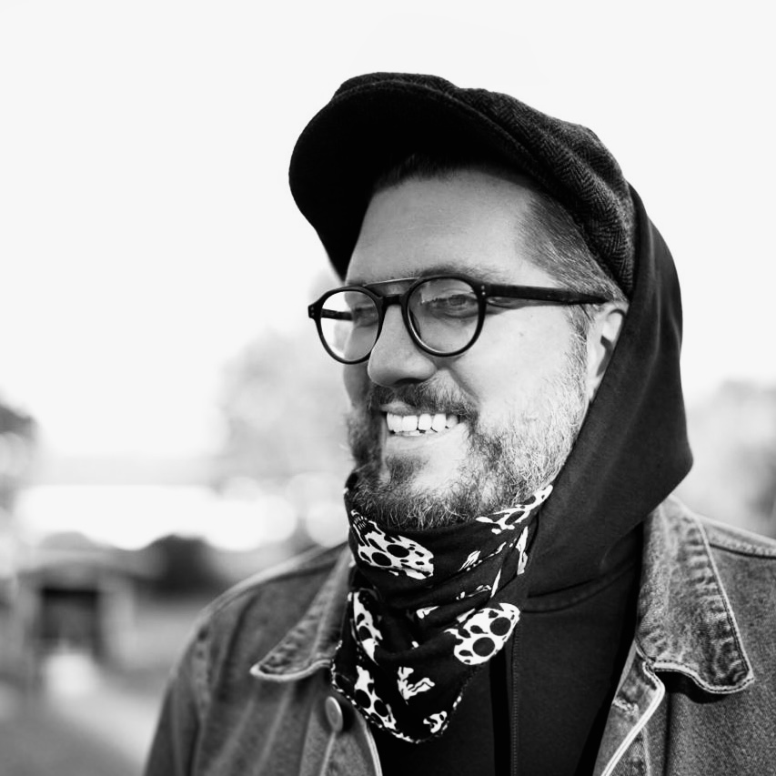 Timo Blume, lachend mit Brille und Schiebermütze, in Schwarzweiß.
